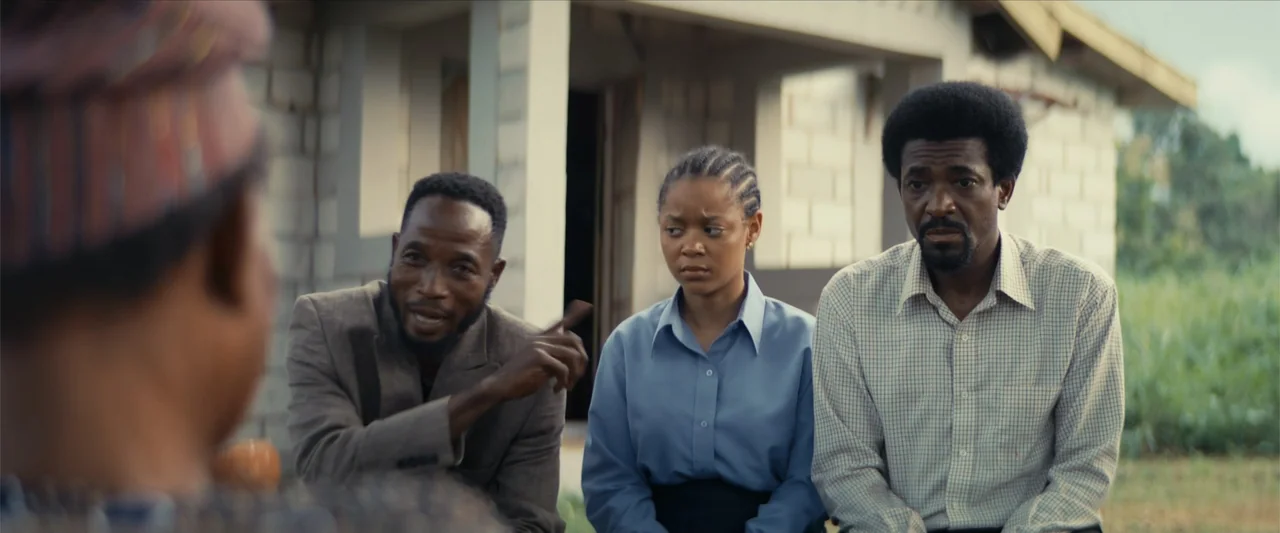 Ms. Kanyin (2025) movie still: medium — Three individuals sit in front of an unfinished concrete block structure outdoors. The man on the left…