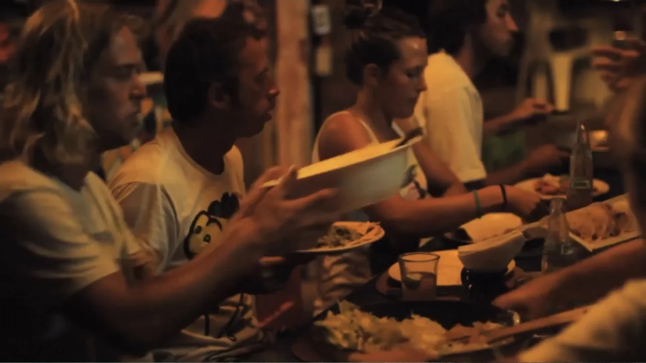 El sueño (2020) movie still: medium — A dimly lit indoor dining area contains a wooden table with food items. Four young adults eat together in a…