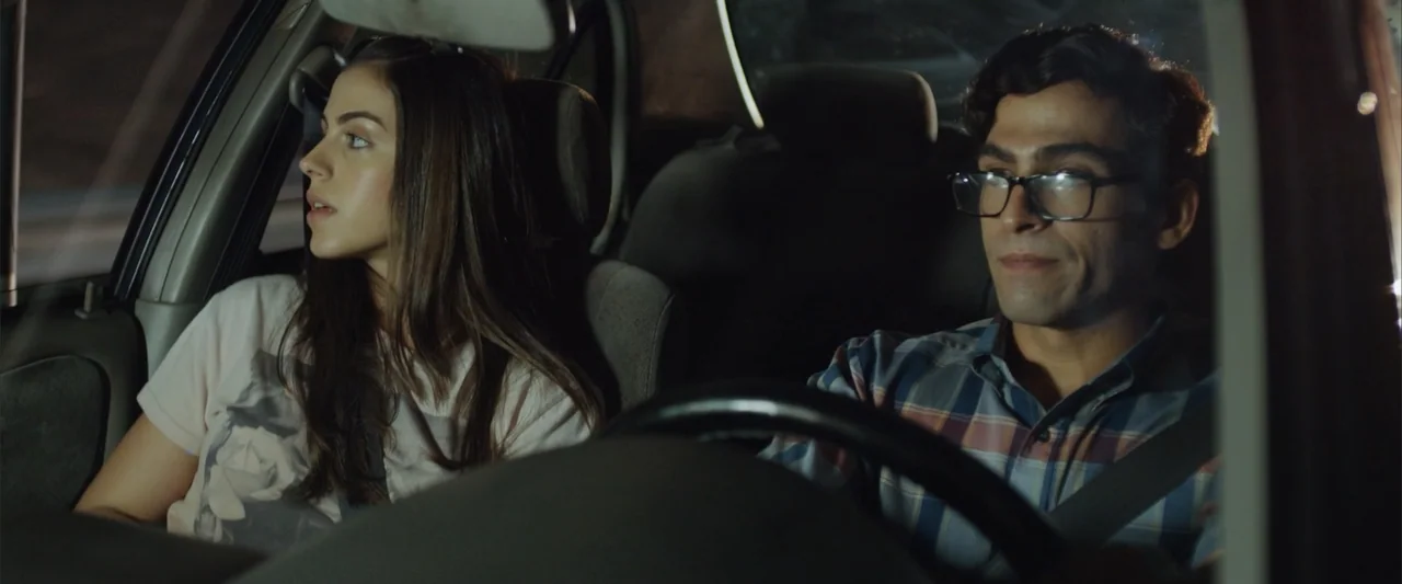 La Musiquita por Dentro (2019) movie still: medium — A car interior at night with a man driving and a woman sitting passenger. They sit side by side…