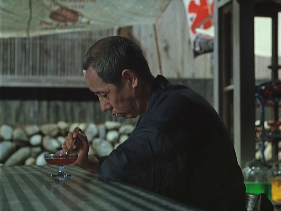 Floating Weeds (1959) movie still: medium — A man sits at a striped table inside a simple, open-sided shop structure. He holds a metal spoon while…