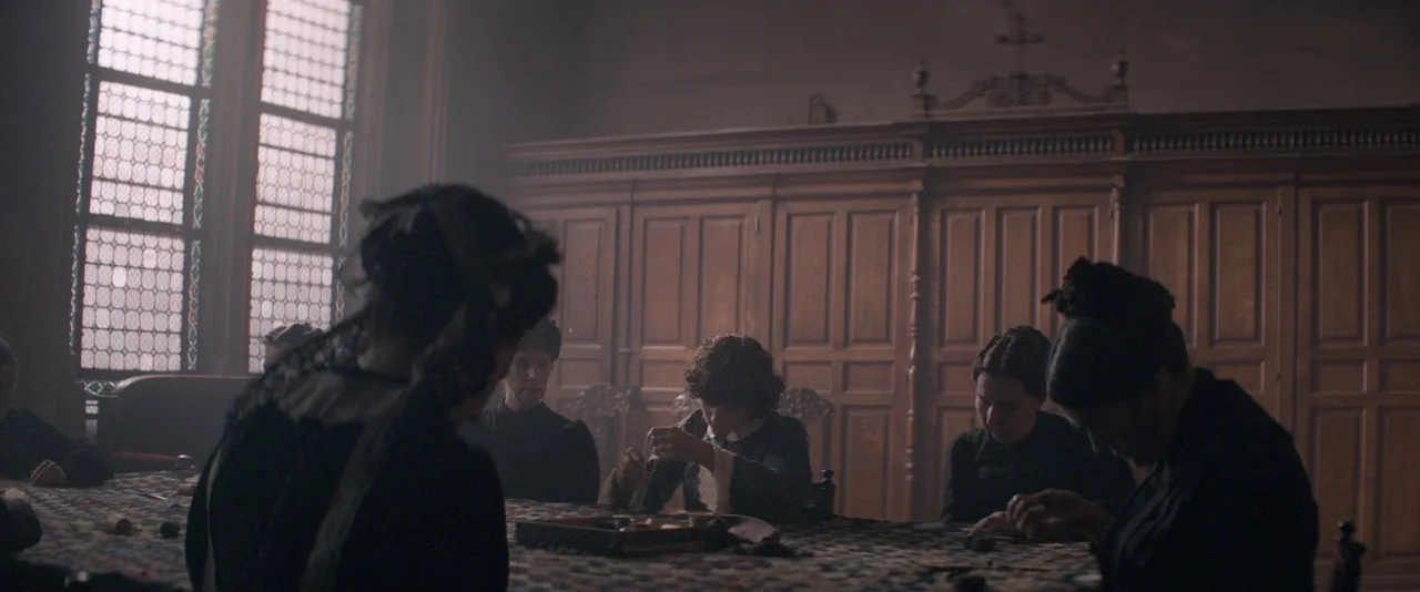 The Dancer movie still: medium — An interior room featuring paneled wooden walls and tall window panes with grid patterns. Several individuals sit…