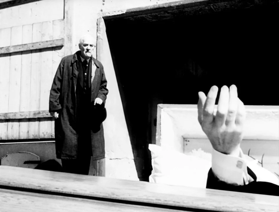 Wild Strawberries (1957) movie still: medium — An outdoor setting with a wooden structure background and a partially open coffin foreground. An…