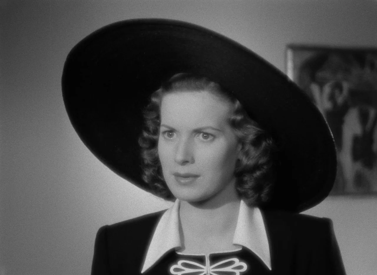 Dance, Girl, Dance (1940) movie still: closeup — A woman stands indoors in front of a plain wall with a framed picture. She wears a large dark hat…