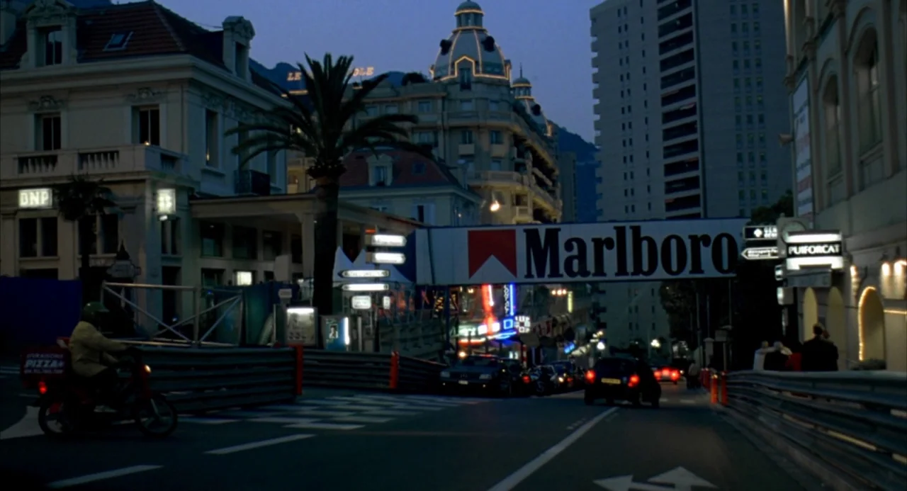 The Good Thief (2002) movie still: wide — An urban street in Monaco at twilight, bordered by tall buildings and race barriers. A large overhead…