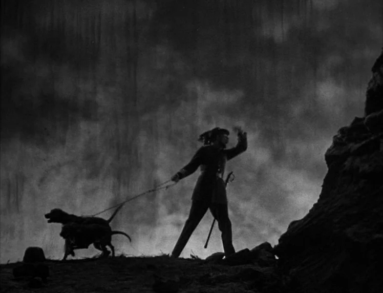 Frankenstein movie still: wide — A soldier leads two dogs across a rugged, desolate rocky landscape against dark clouds. The figure stands in stark…