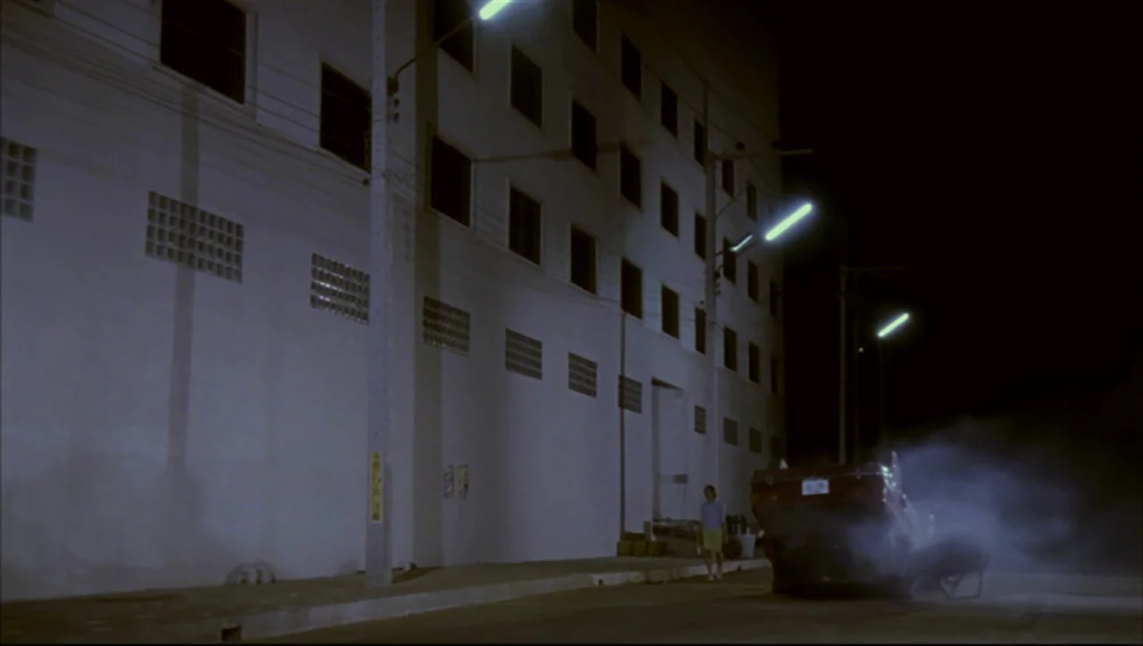 Wild Zero (1999) movie still: wide — A dark city street at night features a large, stark white administrative building wall. A smoking, crashed…