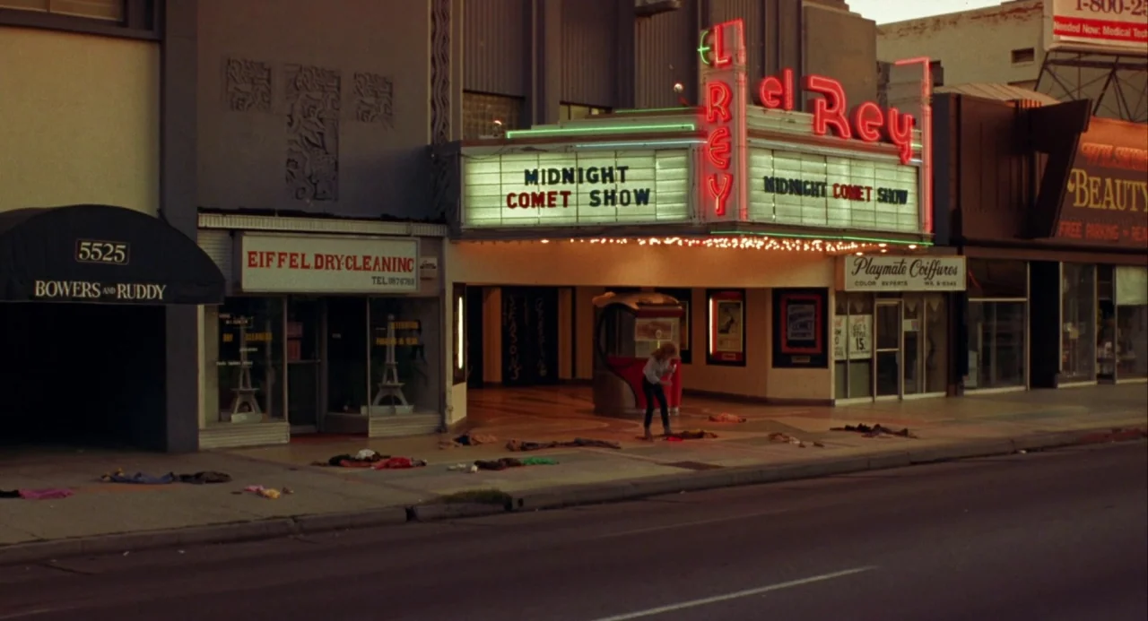 Night of the Comet movie still: wide — A city street at night features a theater marquee with neon lights. A lone woman stands on the sidewalk…