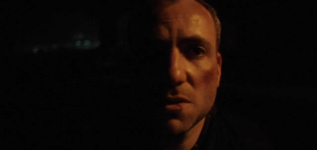 Pusher (1996) movie still: closeup — A dark vehicle interior with blurred distant city lights visible through a window. A man sits in the darkness…
