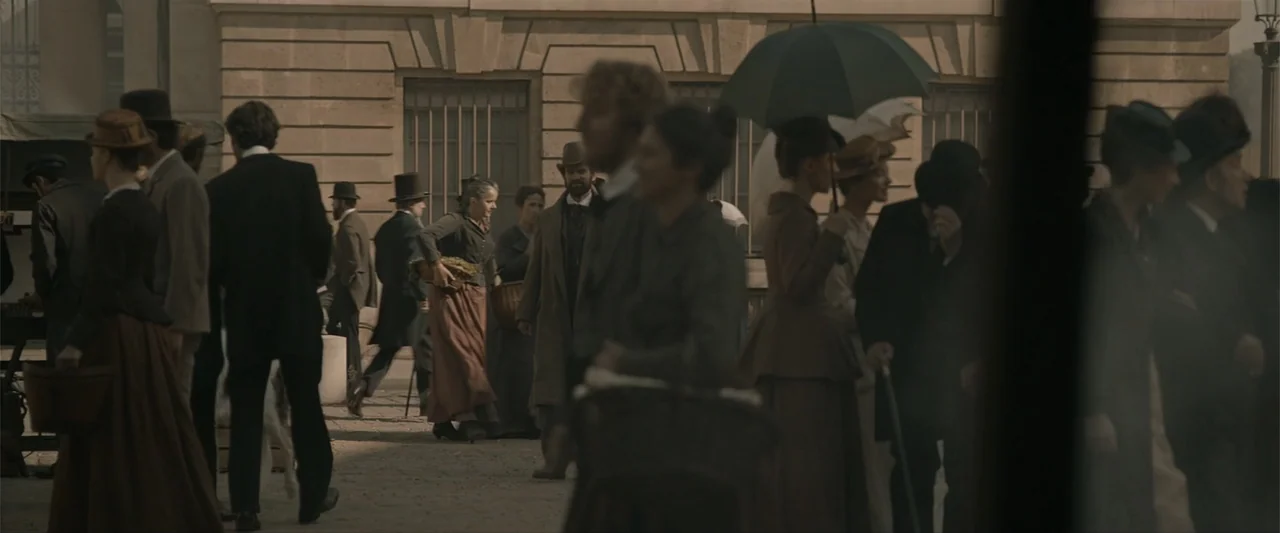 Eiffel (2021) movie still: wide — A 1900s stone plaza with classic architecture and metal-grated windows in the background. A crowd of people in…