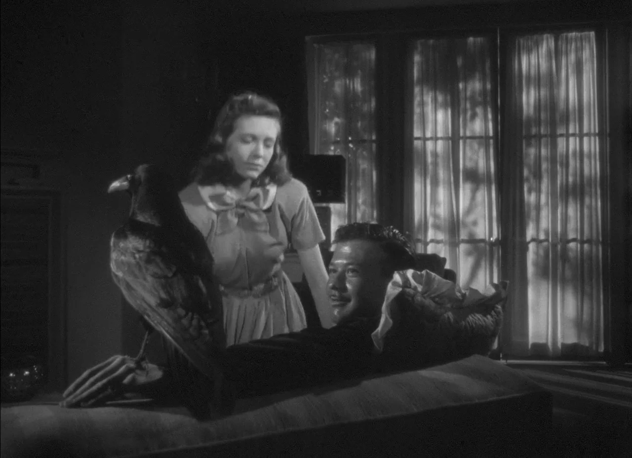 The Amazing Mr. X (1948) movie still: medium — A dark, dimly lit room contains a sofa positioned in front of a window. A woman stands behind the…