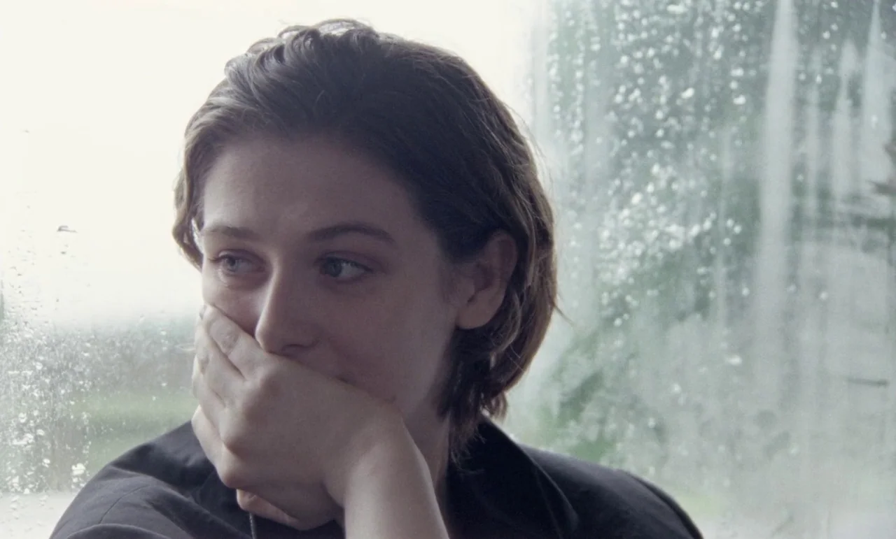 The Souvenir: Part II (2021) movie still: closeup — A close up view of a person seated beside a rain-streaked window pane. The subject leans their…
