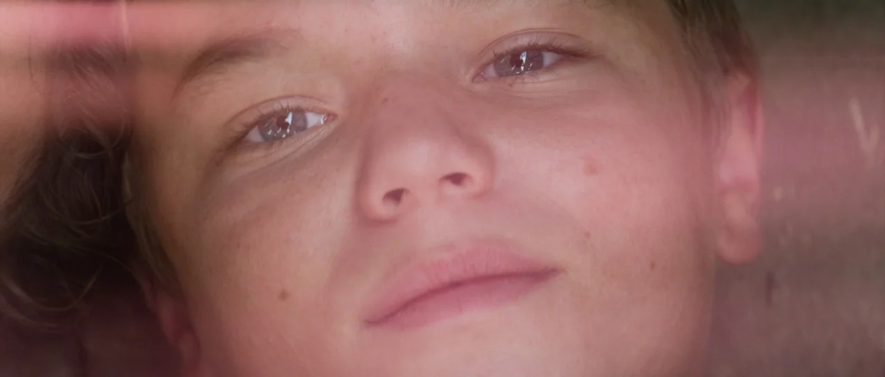 Elysium (2013) movie still: closeup — A close up of a teenage boy inside a vehicle with lens flare. The boy looks directly at the camera while lying…
