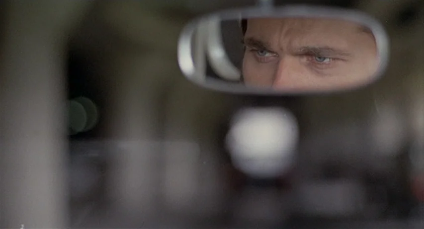The Fifth Cord (1971) movie still: closeup — A man's face is seen reflected in a car's rearview mirror. The rest of the frame is blurred interior…
