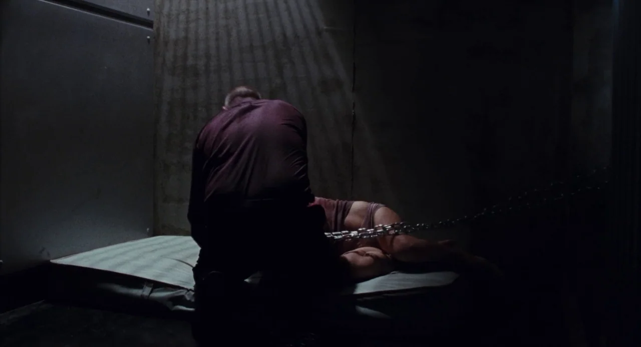 Martyrs (2008) movie still: medium — A dark, spartan room features a single thin mattress placed against a concrete wall. A person kneels over…