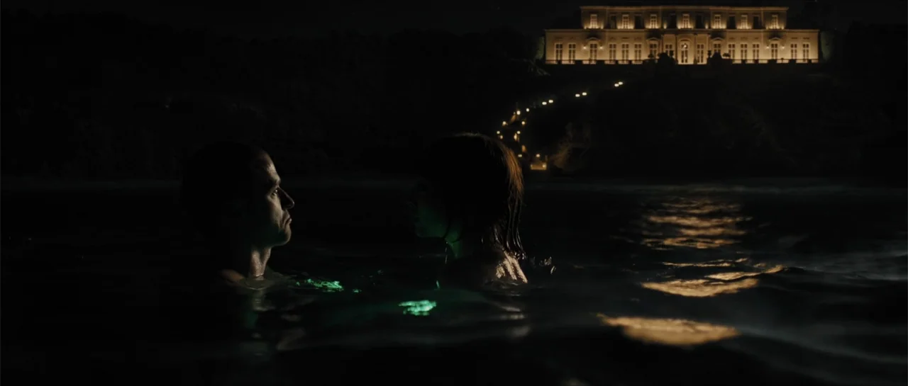 In Time (2011) movie still: medium — A dark ocean at night with a large illuminated mansion on a distant cliff. A man and woman tread water in the…