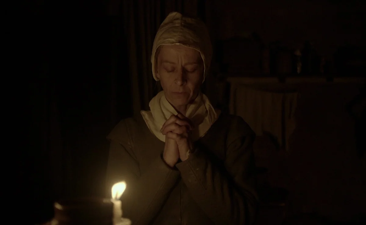 The Witch (2015) movie still: medium — A dimly lit room with simple wooden walls and an obscured background. An older woman with a white bonnet…