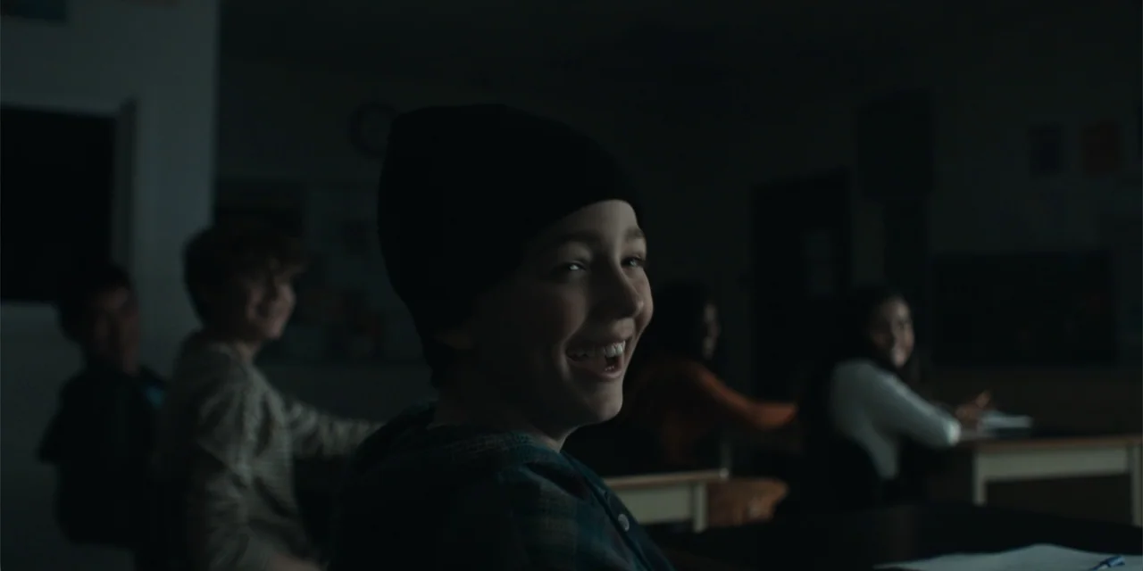 The Monkey (2025) movie still: medium — A dimly lit classroom features students sitting at desks in the background. A young boy wearing a black…