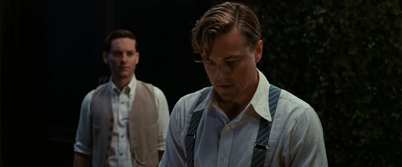 The Great Gatsby (2013) movie still: medium — A dimly lit room with dark paneling and blurred foliage in the background. Two men in vintage 1920s…