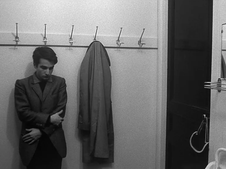Masculin Féminin (1966) movie still: medium — A spartan room features a wooden coat rack mounted on a white wall. A man stands beside a trench coat…