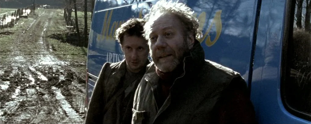 Calvaire (2004) movie still: medium — A muddy rural track runs alongside a blue cargo van on a cloudy day. Two men stand together, facing slightly…