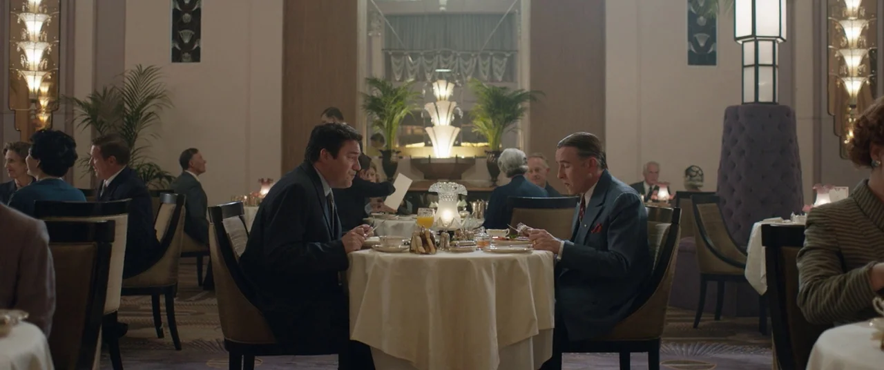 Domino (2005) movie still: wide — A spacious, elegant 1960s hotel dining room features Art Deco mirrors and tall plants. Two men sit at a central…