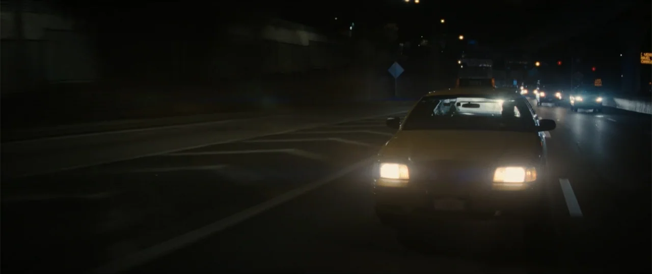 Daddio (2024) movie still: wide — An asphalt highway stretches into the dark distance under dim overhead lighting. A yellow sedan occupies the right…