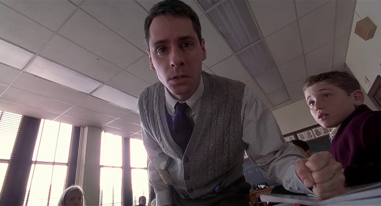 The Sixth Sense (1999) movie still: medium — A school classroom with fluorescent lights and windows covered by vertical blinds. A male teacher leans…