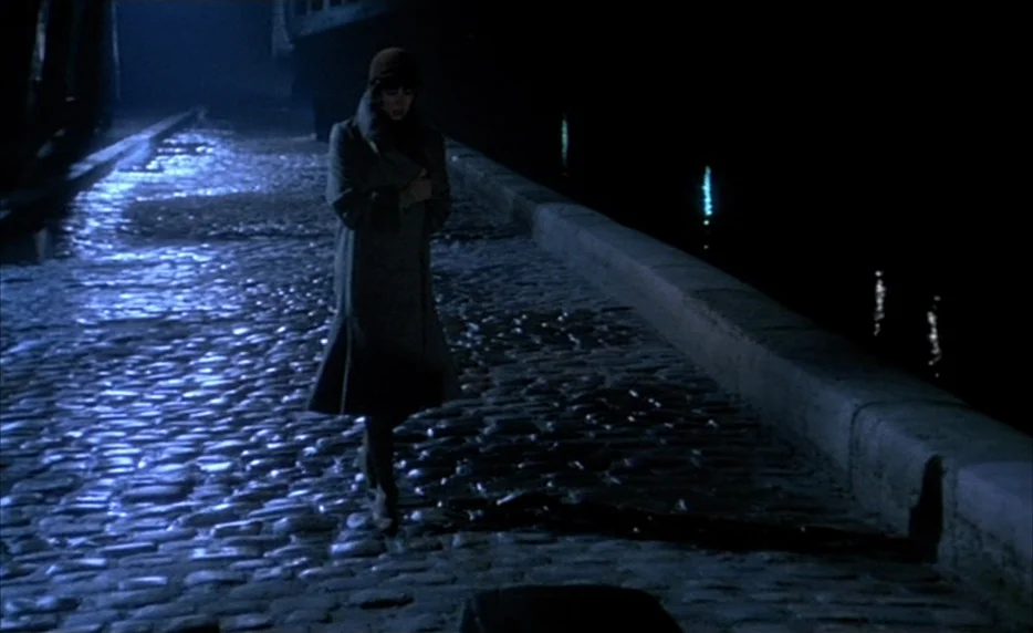 Mélo (1986) movie still: fullbody — A dark, wet cobblestone quay borders a body of water at night. A woman in a long coat walks alone along the…