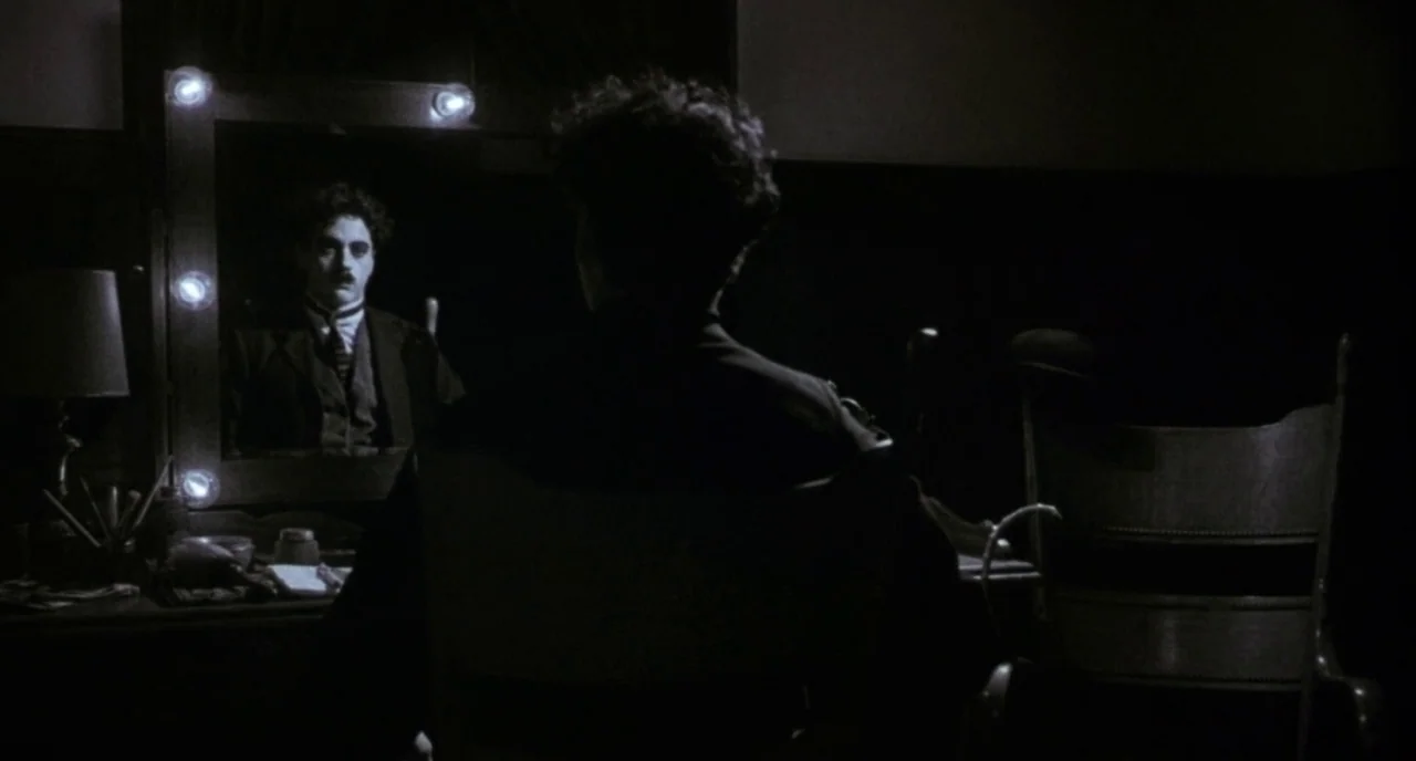 Chaplin (1992) movie still: medium — A dimly lit dressing room contains a large vanity with illuminated light bulbs. A man in a suit sits before the…