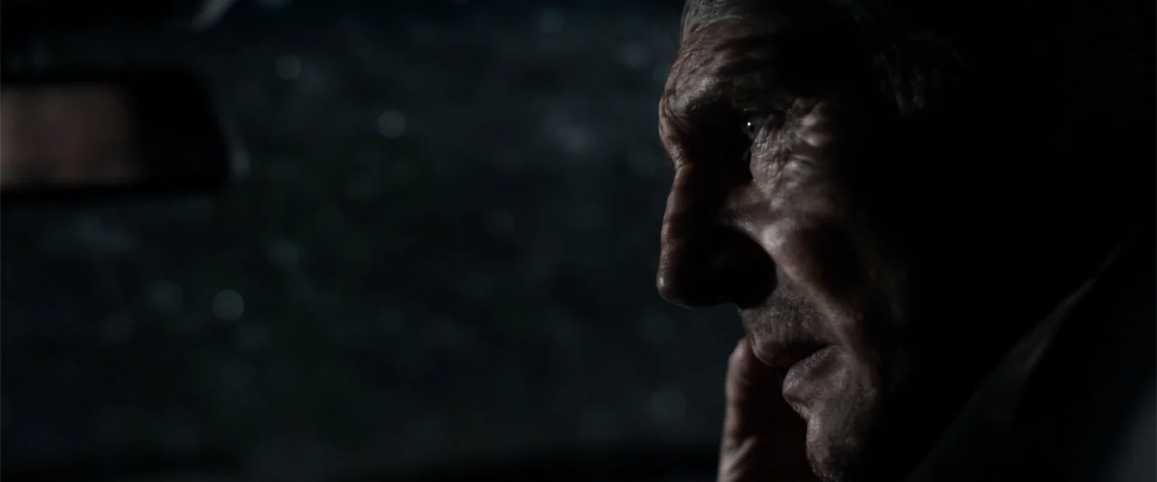 A Walk Among the Tombstones (2014) movie still: closeup — The interior of a dark car at night with a rain-slicked window visible. An elderly man…