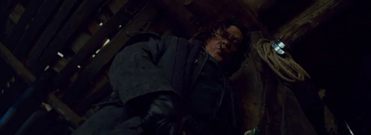 The Hateful Eight (2015) movie still: medium — A dark, wooden interior with textured logs and a rope hanging on the side. A man stands looking down…