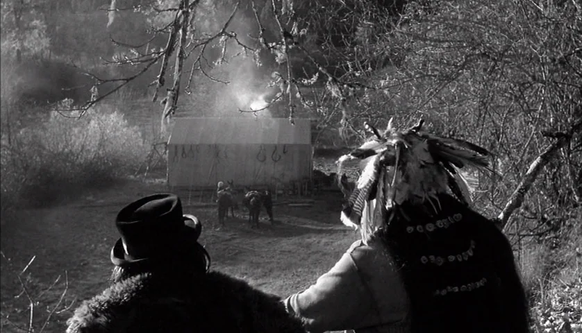 Dead Man (1995) movie still: wide — A clearing in a dense woodland features a canvas tent with hanging objects. Two men stand on a ridge looking…