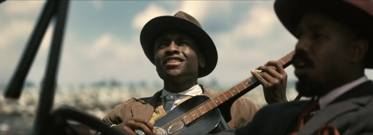 Sinners (2025) movie still: medium — Two men sit in a convertible car traveling along a road during the day. The men play guitar and sing while…