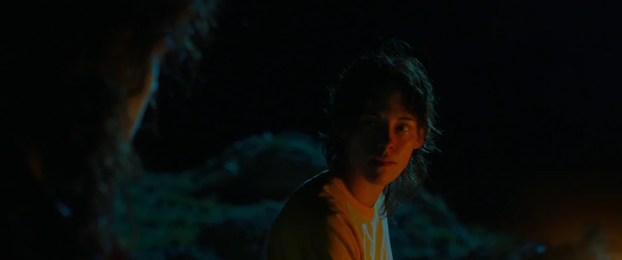 Love Lies Bleeding (2024) movie still: medium — A dark outdoor space at night with a person illuminated by firelight. A woman with short hair looks…