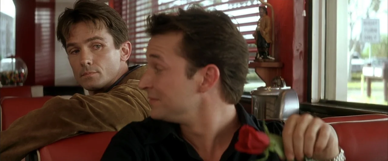 Enough (2002) movie still: medium — A 1990s diner booth with red vinyl seating and venetian blinds on windows. Two men sit together in the booth as…