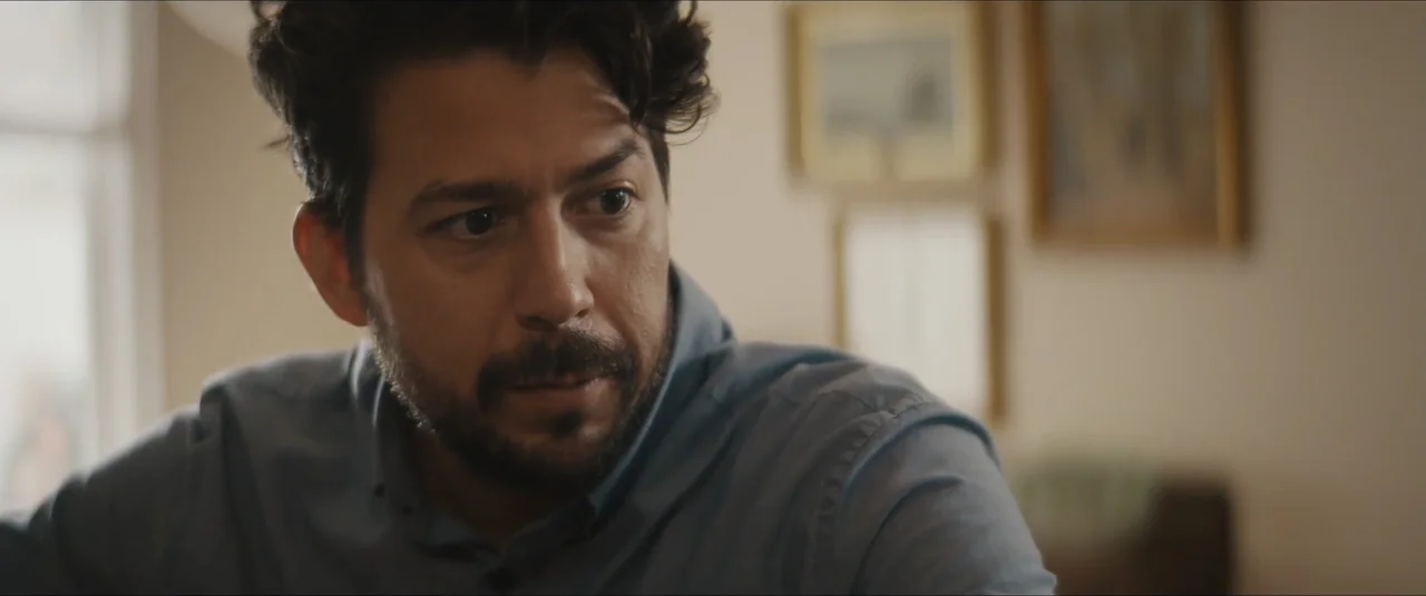 Stalker (2020) movie still: closeup — A residential room features light walls decorated with framed prints. A man stands in the foreground looking…