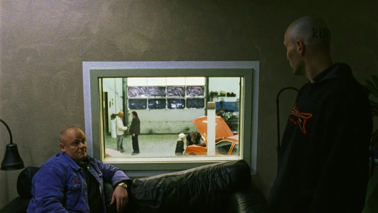 Pusher II (2004) movie still: medium — An interior office space with a large window looking into a vehicle repair area. A man sitting on a leather…