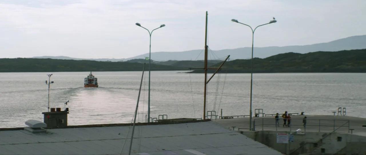 Silence (2012) movie still: wide — A coastal pier extends into a calm bay under a bright, overcast sky. A small boat moves away from the land while…