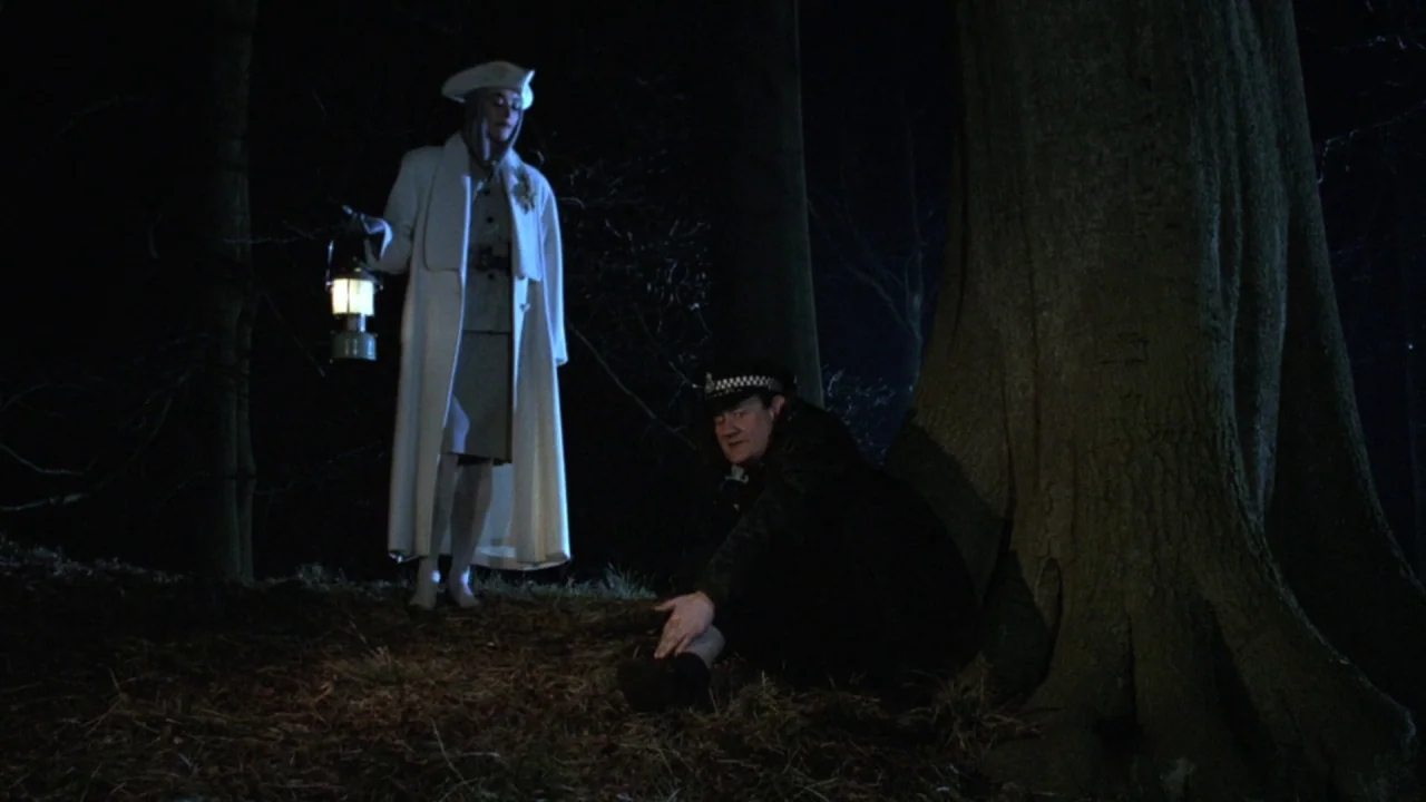 The Lair of the White Worm (1988) movie still: fullbody — A dark, wooded environment at night featuring tall trees and leaf-covered ground. A woman…