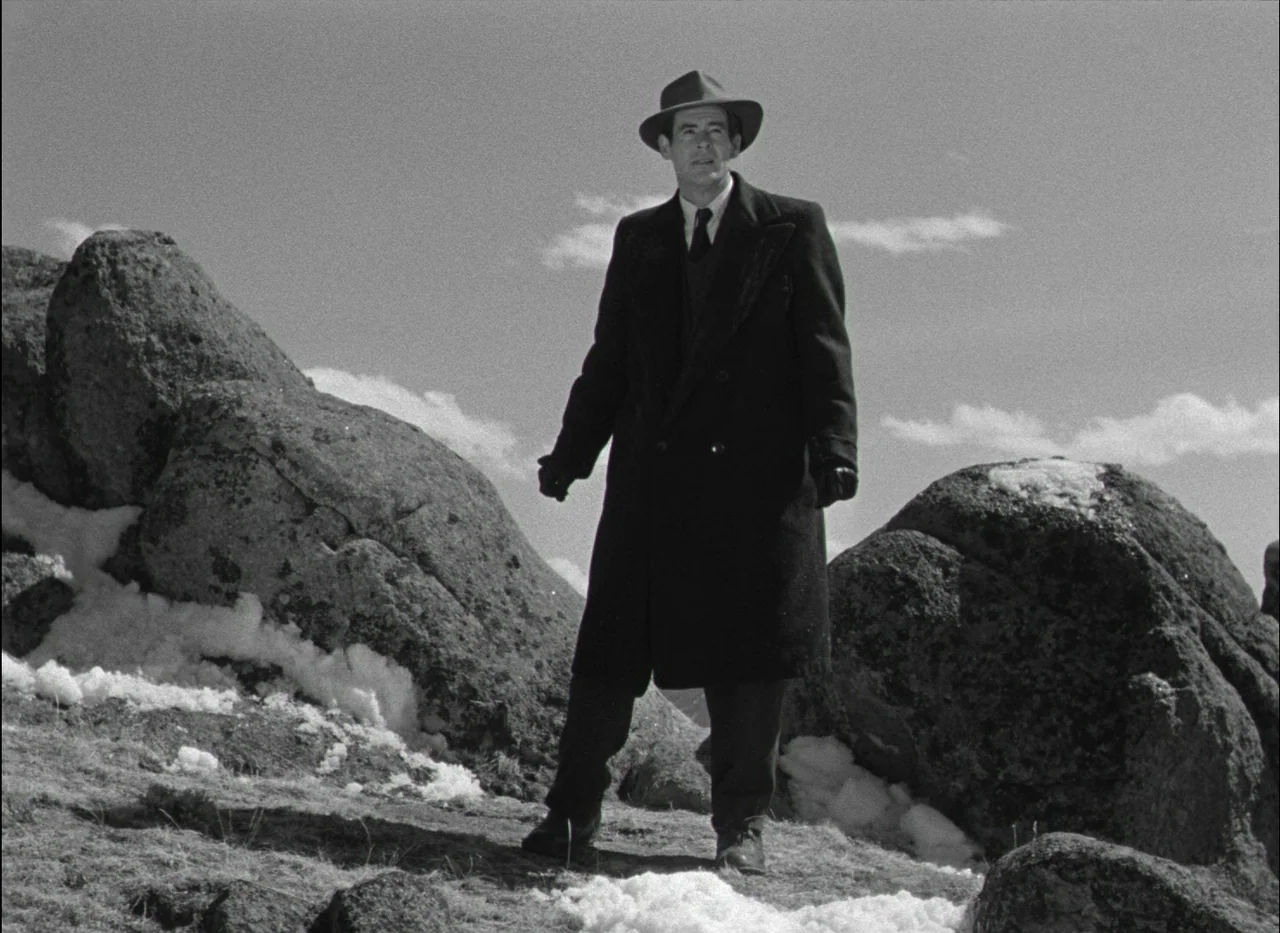 On Dangerous Ground (1951) movie still: fullbody — A man stands on a rugged, snow-dusted hillside under a bright sky. Large rock formations flank…