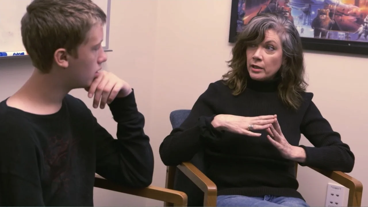 A Dangerous Son (2018) movie still: medium — An office interior features a white wall and a framed poster on the wall. A woman gestures while…
