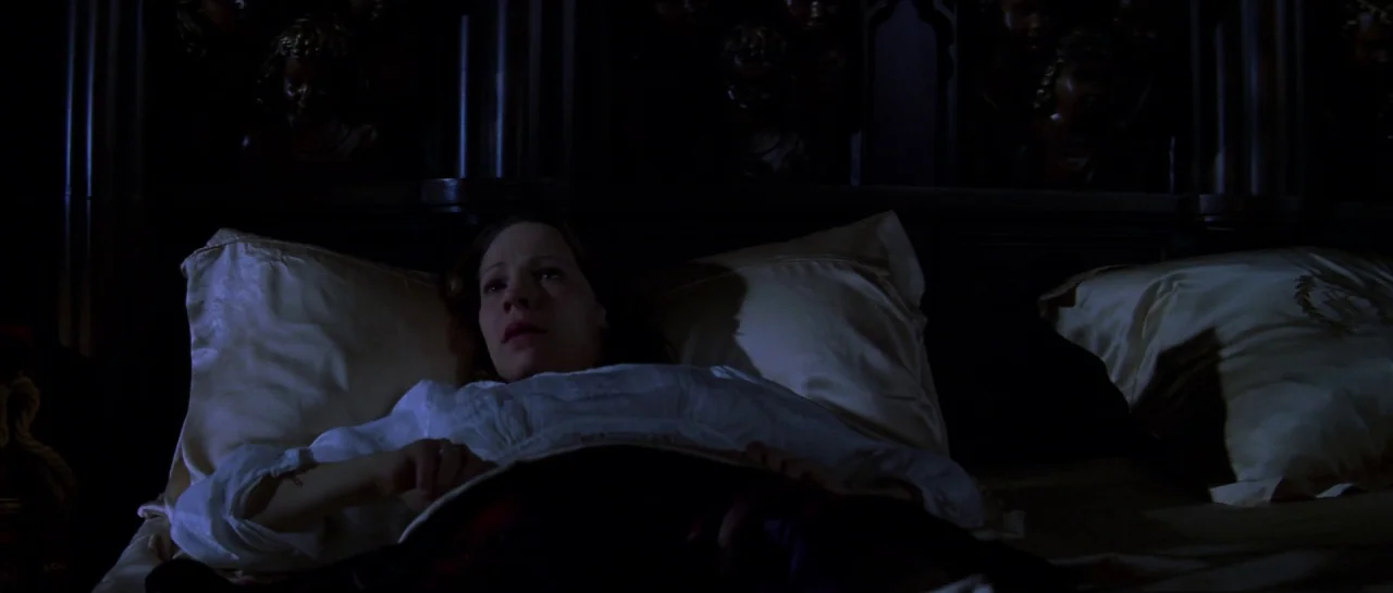 The Haunting (1999) movie still: medium — A dark bedroom with an ornate dark wood headboard carved with numerous small figures. A woman lies flat in…