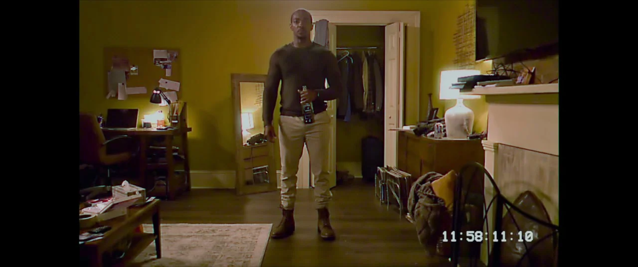 Synchronic (2019) movie still: fullbody — A furnished interior space with mustard-yellow walls features a desk, a mirror, and a fireplace. A man in…