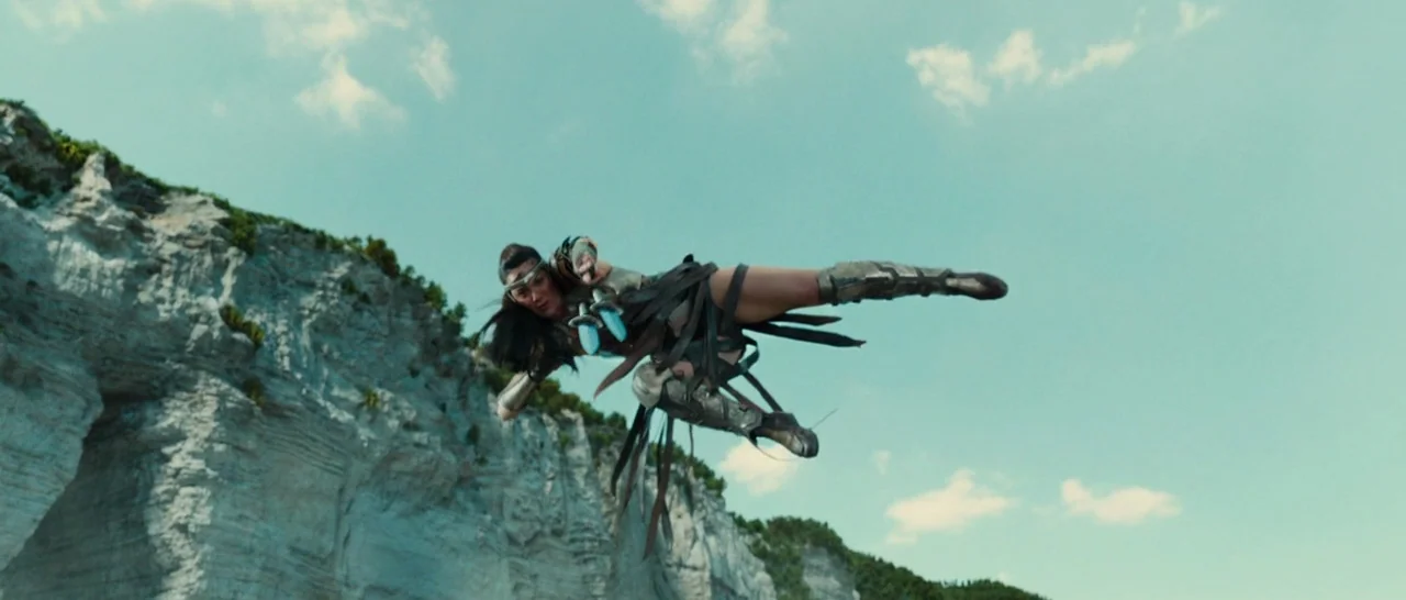 Wonder Woman (2017) movie still: fullbody — A sheer white limestone cliff face sits against a clear blue daytime sky. A female warrior in metallic…