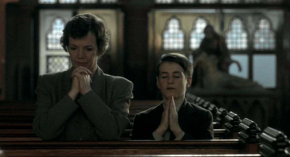 The Long Day Closes (1992) movie still: medium — A woman and a young man sit together in a wooden pew inside a church. Both have their eyes closed…