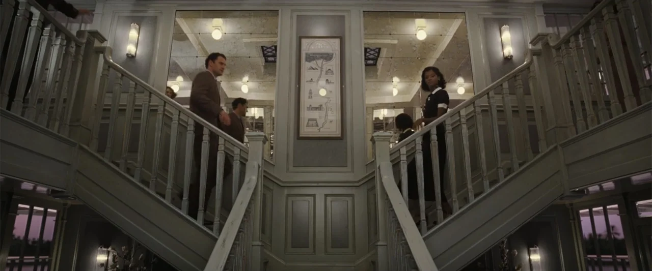 Death on the Nile (2022) movie still: medium — A symmetrical grand staircase features wooden railings and a framed map on the wall. A man in a brown…
