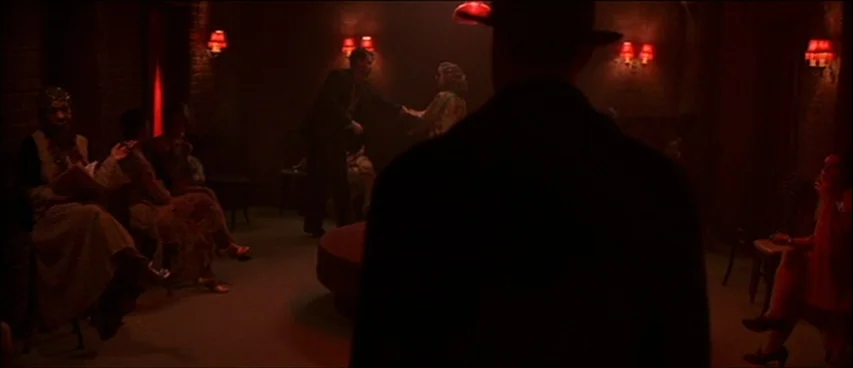 Road To Perdition (2002) movie still: medium — A dark, dimly lit lounge space features brick walls and red wall sconces. A lone figure in the…
