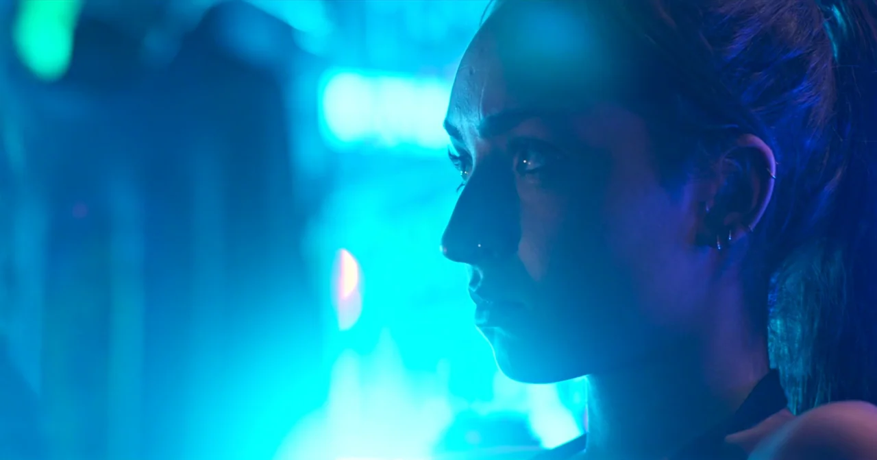 T Blockers (2023) movie still: closeup — A city street at night illuminated by intense blue neon signage in the background. A woman in a dark top…