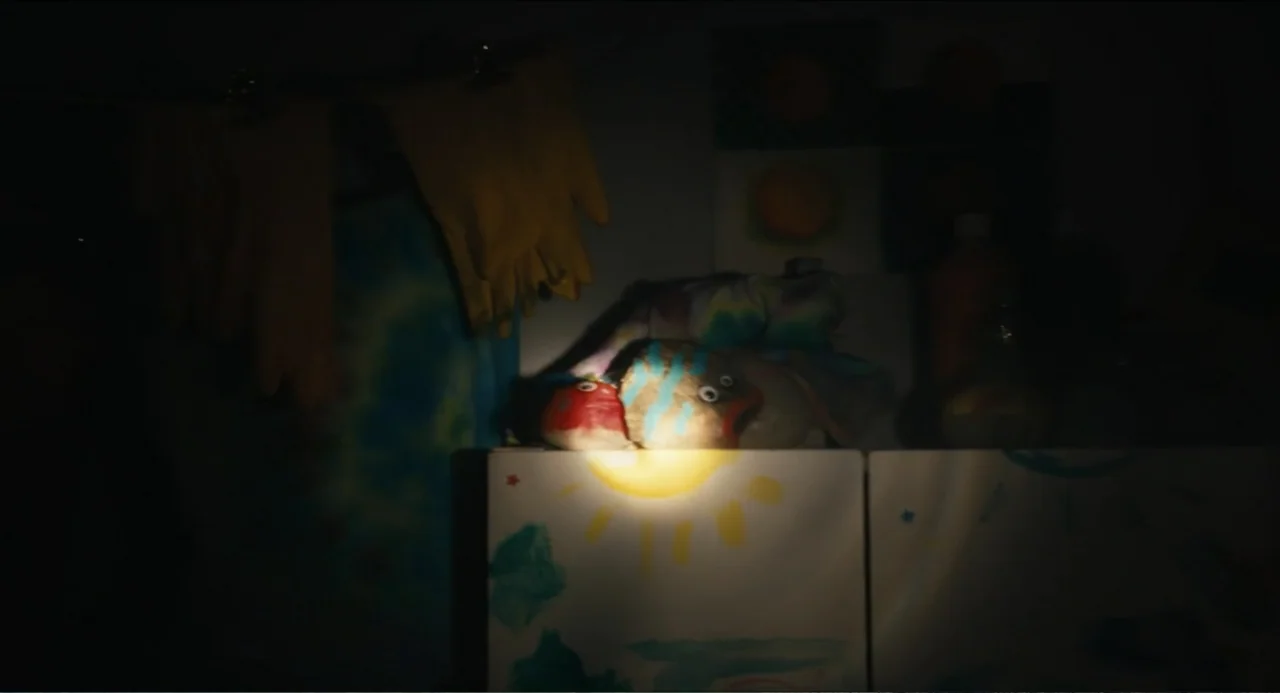 Hell of a Summer (2025) movie still: medium — A dark indoor space contains shelves decorated with simple child-like painted suns. Two stuffed toys…
