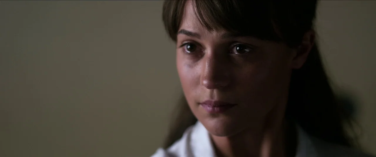 Earthquake Bird movie still: closeup — A dimly lit room with a plain, neutral-toned wall in the background. A woman in a white collared shirt stares…